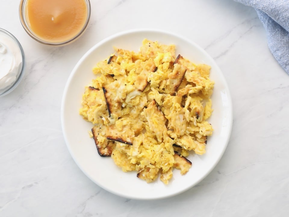 Overhead horizontal shot - a white plate with matzo brei eggs and matzo scrambled, small dishes of applesauce and sour cream beside, with a blue towel in background, on a white marble counter.