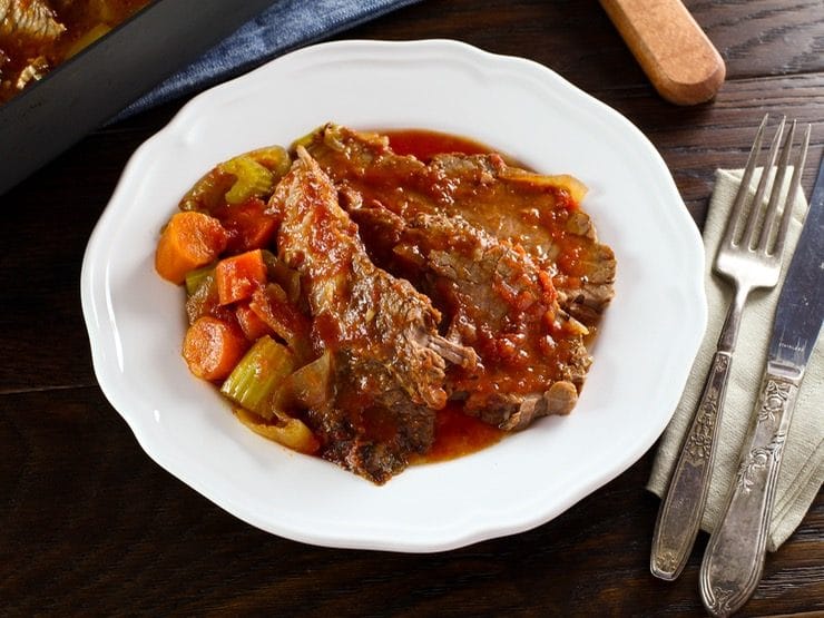 Slices of Holiday Brisket with red sauce, cooked carrots and celery on white plate with fork, knife and cloth napkin, roasting pan in background on wooden surface.