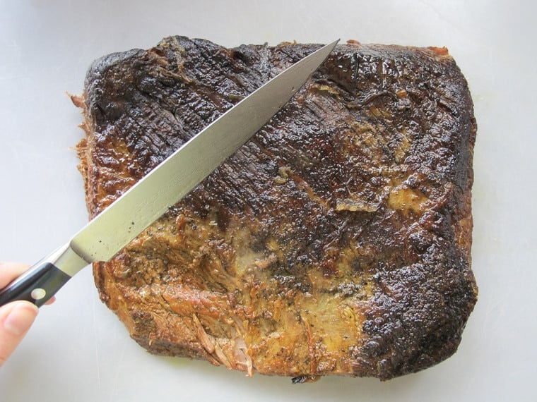 Cooked brisket on white cutting board, hand holding knife up against the grain.