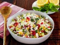 Square crop - bowl filled with fresh Israeli Salad with Feta, tomatoes, cucumbers and bell peppers. Mint and lemon in the background, cloth napkin and wooden spoon beside the bowl.