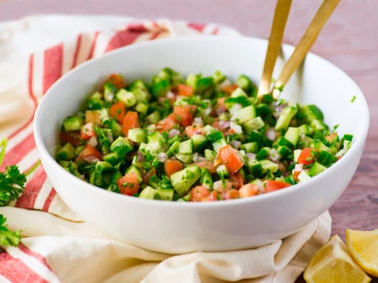 Bowl of Israeli Salad with serving forks on wooden table with lemon slices on the side.