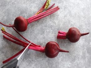 Three beets, with stems being trimmed by kitchen shears, on grey concrete background.