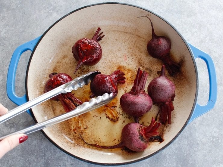 Flipping half-cooked beets in enameled casserole dish with stainless tongs on a concrete background.