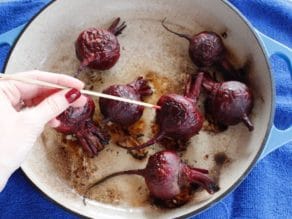 Hand skewering a roasted beet with a wooden skewer in an enameled cast iron casserole dish on a blue towel.