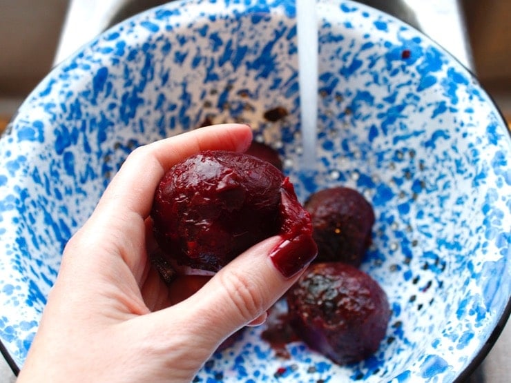 Hand peeling roasted beet over a blue speckled colander with running water in background.