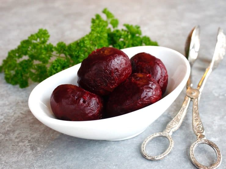 Roasted peeled beets in a white bowl with fresh parsley and antique silver tongs.