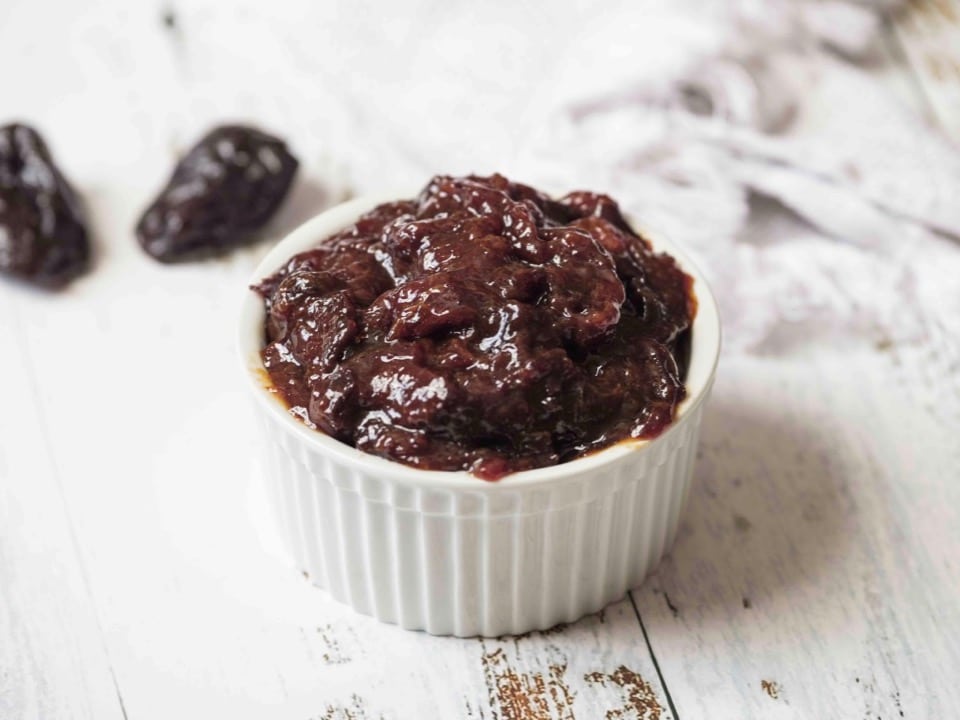 Horizontal shot - prune filling in a white ramekin on white wood background with prune in background.