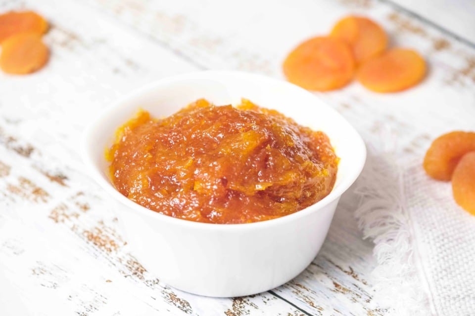 Horizontal Shot - Apricot lekvar - apricot butter for cookie and pastry filling in a small white dish on a rustic white wood background, with dried apricots in the background.