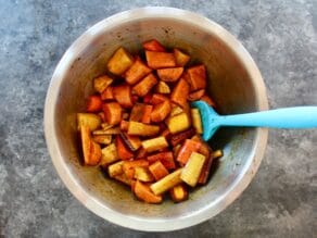 Bowl of root vegetables cut up tossed with molasses and olive oil, blue spatula, on concrete background.