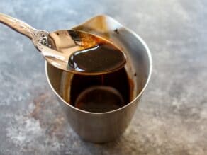 Rich thick balsamic reduction in spoon over measuring cup on concrete background.