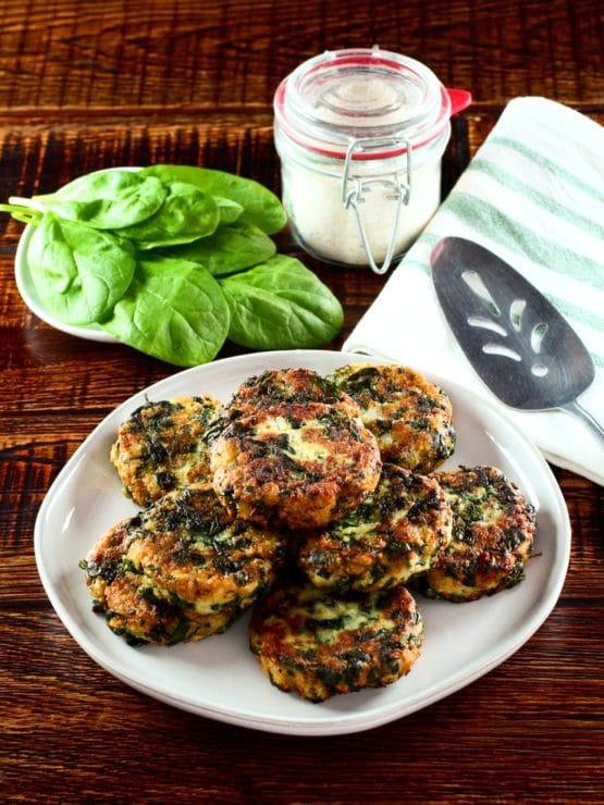 Plate of spinach keftes piled on a white plate with fresh spinach, canister of breadcrumbs, spatula and cloth napkin in background.