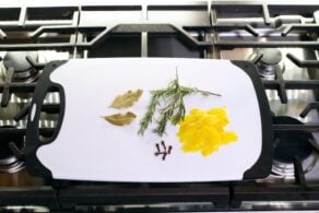 Ingredients for herb bundle on cutting board on stovetop - bay leaves, rosemary, cloves, lemon peel.