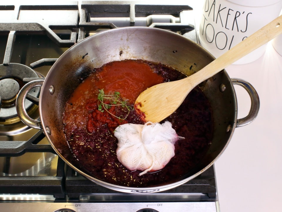 Sauce and herb bundle in pan on stovetop with wooden spoon.