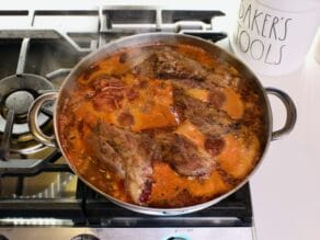 Pan of lamb shanks braising in red wine, tomato paste and liquid on stovetop.