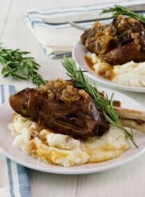 Square featured shot. Tender slow braised lamb shank on mashed potatoes with sprig of rosemary on a white plate, blue and white cloth napkin and fork on white table beneath. Another plate with shank and a sprig of rosemary in background.