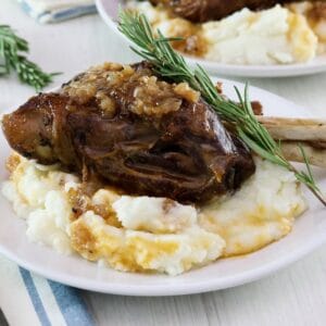 Frontal horizontal shot. Tender slow braised lamb shank on mashed potatoes with sprig of rosemary on a white plate, blue and white cloth napkin and fork on white table beneath. Another plate with shank and a sprig of rosemary in background.