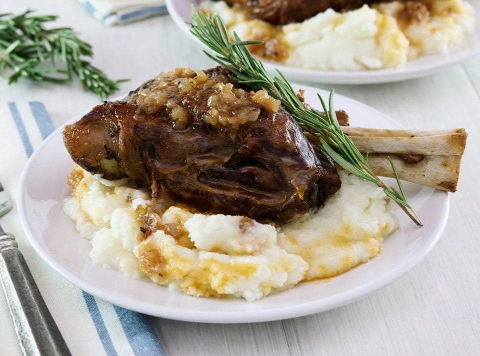 Frontal horizontal shot. Tender slow braised lamb shank on mashed potatoes with sprig of rosemary on a white plate, blue and white cloth napkin and fork on white table beneath. Another plate with shank and a sprig of rosemary in background.