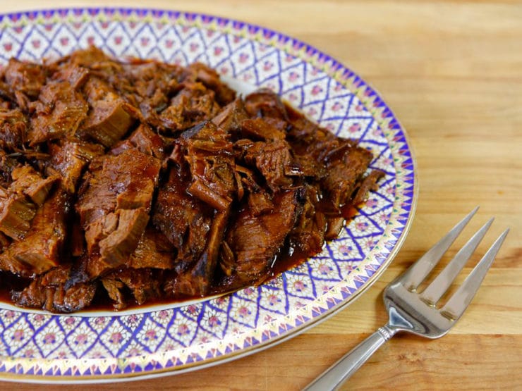 Slow Cooker Honey Barbecue Brisket pieces on decorative plate on wooden cutting board with serving fork.