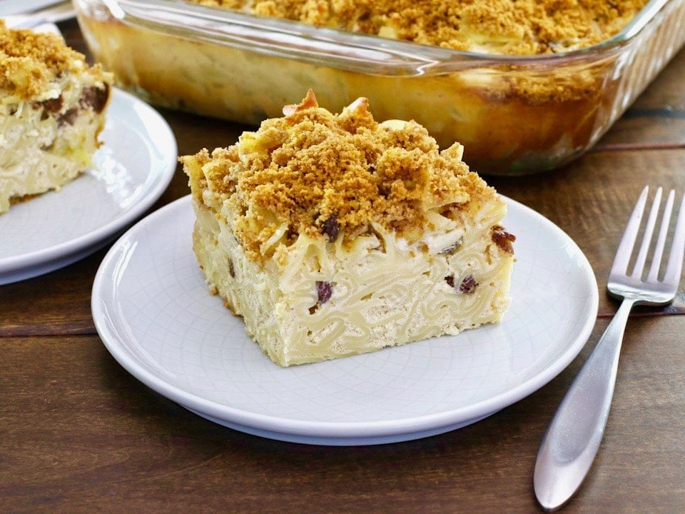 Horizontal Crop - close up of a slice of creamy noodle kugel with raisins and pineapples, with fork beside it, on wooden table. Baking dish of kugel and another slice in background.
