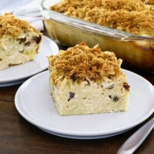 Horizontal Crop - close up of a slice of creamy noodle kugel with raisins and pineapples, with fork beside it, on wooden table. Baking dish of kugel and another slice in background.