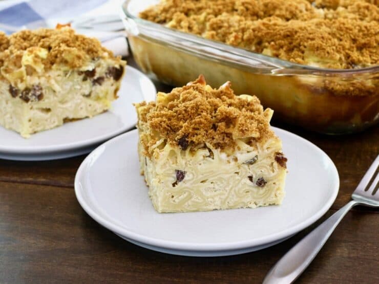Horizontal Crop - close up of a slice of creamy noodle kugel with raisins and pineapples, with fork beside it, on wooden table. Baking dish of kugel and another slice in background.