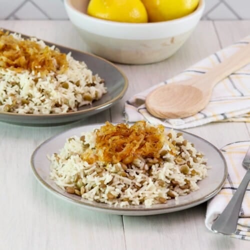 Horizontal shot. In foreground, small dish of mujadara lentils and rice topped with caramelized onions, with fork and cloth napkin beside it. In background, oblong platter of mujadara topped with caramelized onions, spoon and cloth napkin beside it; dish of three lemons in background, against a tile wall.