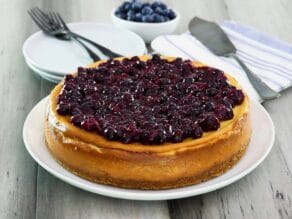 Cheesecake whole on plate, topped with thick rich blueberry topping with fresh blueberries in background. Utensils and plates behind.