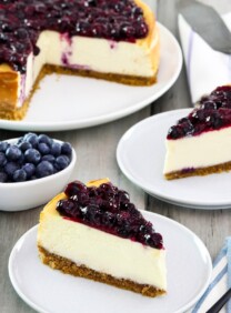 Square crop - two slices of cheesecake on plates with fresh blueberries beside them, a whole cheesecake with slices cut out and pie server in background.