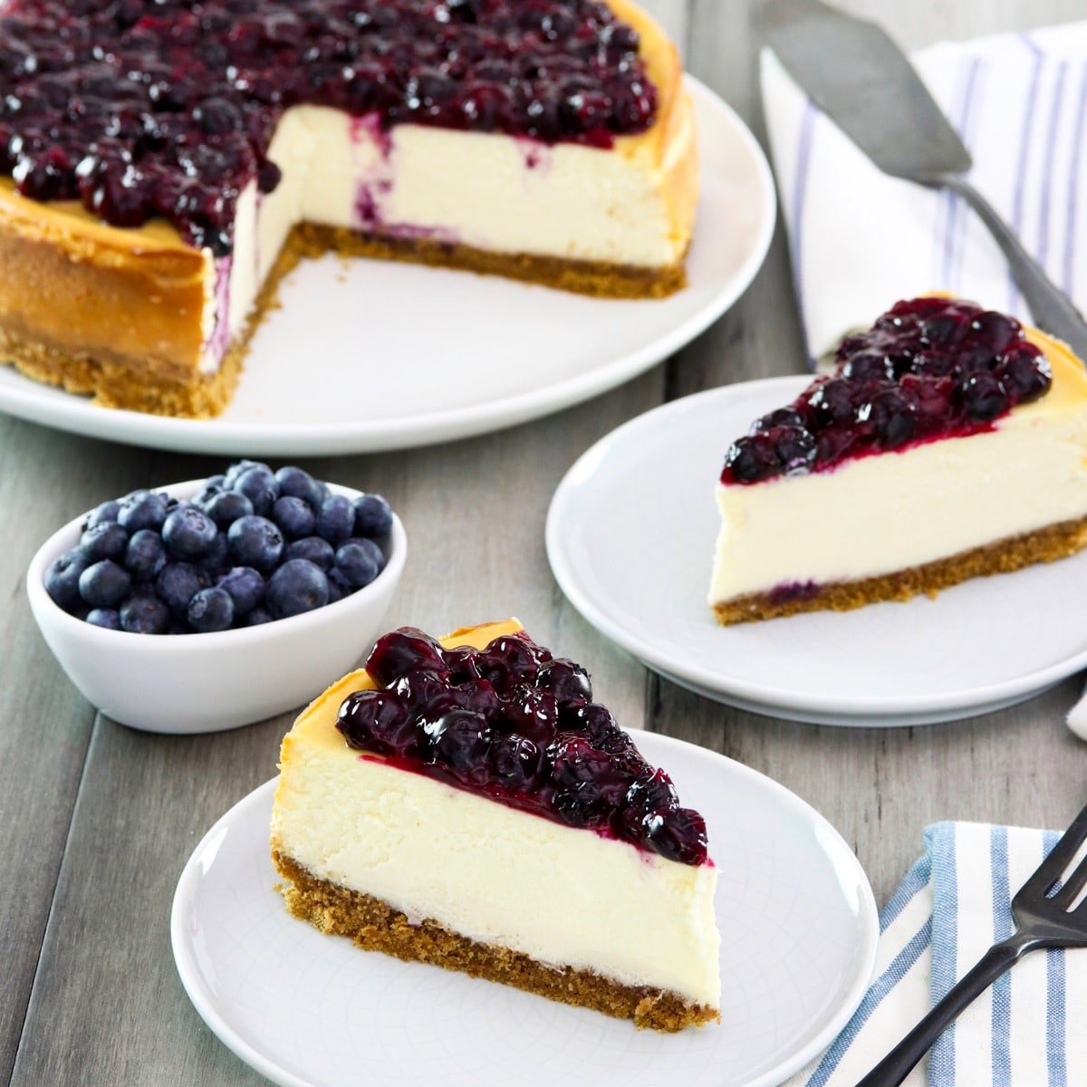 Square crop - two slices of cheesecake on plates with fresh blueberries beside them, a whole cheesecake with slices cut out and pie server in background.