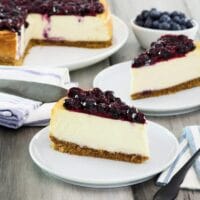Horizontal crop - two slices of cheesecake on plates with fresh blueberries beside them, a whole cheesecake with slices cut out and pie server in background.