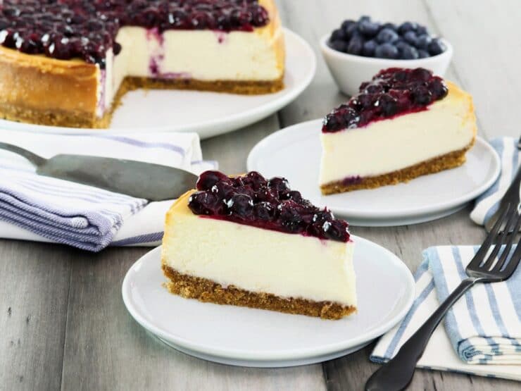 Horizontal crop - two slices of cheesecake on plates with fresh blueberries beside them, a whole cheesecake with slices cut out and pie server in background.