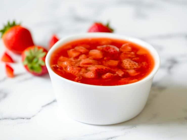 A white dish of strawberry topping on a marble surface, strawberries in background. 