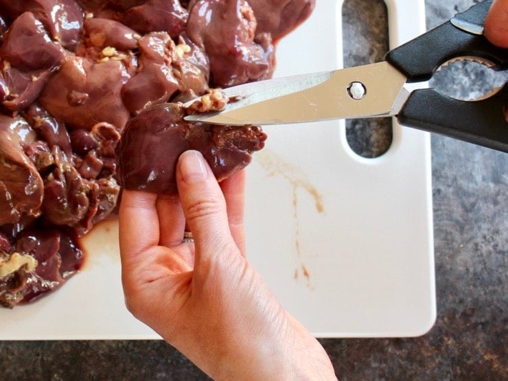 Removing tough tendons from chicken liver with kitchen shears over white cutting board.