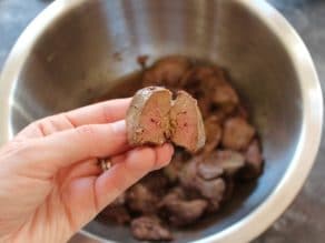 Hand holding chicken liver, cooked, broken in half, to show pinkish interior. Held over a bowl full of cooked chicken livers.