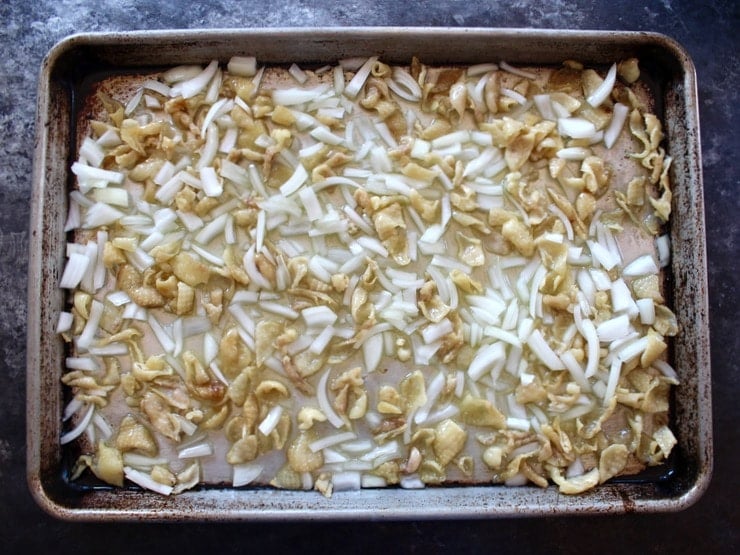 Pieces of chicken skin browning on a baking sheet with raw onion pieces.