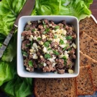 Horizontal shot - dish of chopped liver garnished with chopped hard boiled egg and fresh parsley, with lettuce, serving knife and rye bread pieces, on a wooden cutting board.