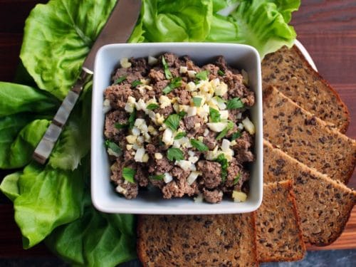 Horizontal shot - dish of chopped liver garnished with chopped hard boiled egg and fresh parsley, with lettuce, serving knife and rye bread pieces, on a wooden cutting board.