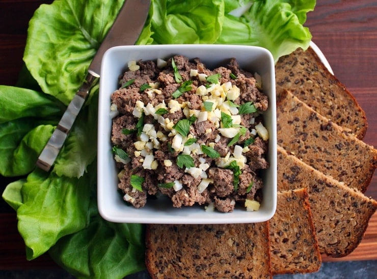 Horizontal shot - dish of chopped liver garnished with chopped hard boiled egg and fresh parsley, with lettuce, serving knife and rye bread pieces, on a wooden cutting board.