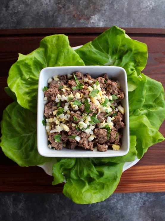 Vertical shot - dish of chopped liver garnished with chopped hard boiled egg and fresh parsley, resting on a bed of lettuce on a wooden cutting board. 