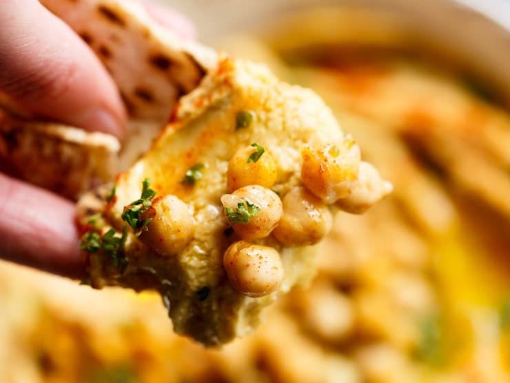 Close up of hand dipping piece of pita bread into hummus with whole chickpeas and fresh parsley garnish.