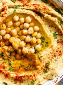 Closeup of hummus in dish topped with paprika, olive oil, parsley and chickpeas, crudités on the side.