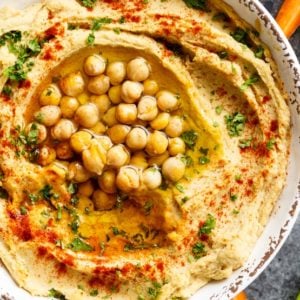 Closeup of hummus in dish topped with paprika, olive oil, parsley and chickpeas, crudités on the side.