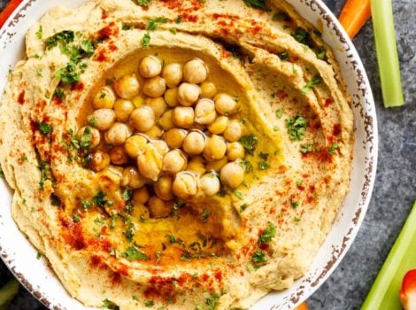 Closeup of hummus in dish topped with paprika, olive oil, parsley and chickpeas, crudités on the side.