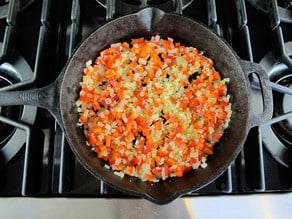 Diced onion and bell pepper sautéing in black cast iron frying pan on stovetop.