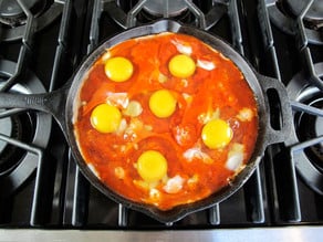 Eggs cooking on top of tomato sauce with pepper and onion sautéing in black cast iron frying pan on stovetop.