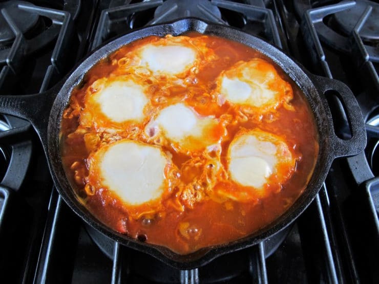 Eggs cooked on tomato sauce sautéing in black cast iron frying pan on stovetop.