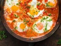 Shakshuka Pinterest Pin - a skillet filled with tomatoes and cooked eggs on top, with a sprinkle of green herbs.