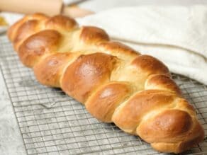 Front view horizontal shot - three strand challah braid fully baked, cooling on wire cooling rack wtih cloth napkin in background.