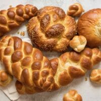 Overhead shot - multiple challah braids - three strand, four strand, round braid, turban challah, and challah rolls, on marble countertop with cloth napkin beneath.