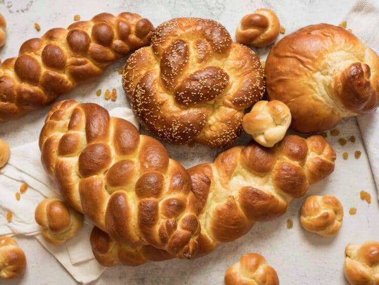 Overhead shot - multiple challah braids - three strand, four strand, round braid, turban challah, and challah rolls, on marble countertop with cloth napkin beneath.